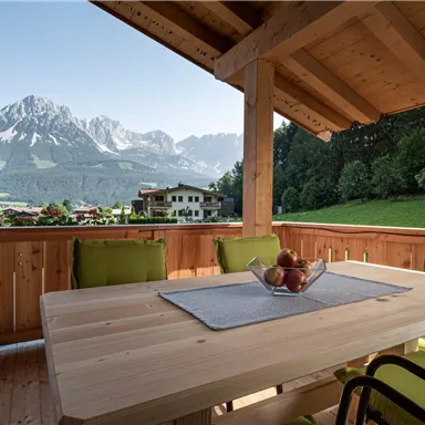 A cozy terrace with a wooden chair and a table decorated with a fruit bowl. In the background, majestic mountains and a green meadow can be seen.