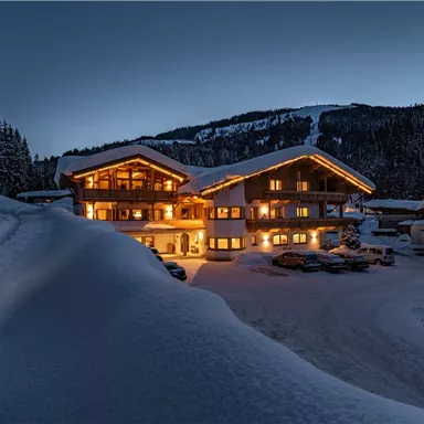 A cozy chalet in the snow, illuminated by warm light. The surroundings are surrounded by trees and mountains.