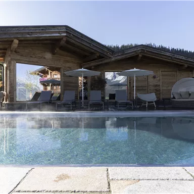 A beautiful outdoor pool with loungers and sun umbrellas. In the background, there is a modern wooden building and the surrounding mountainous landscape.