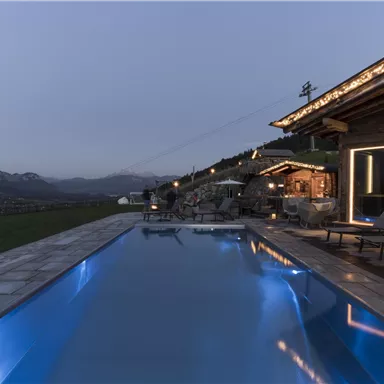 A modern outdoor area with a blue pool and a cozy terrace. In the background, mountains and an evening atmosphere are visible.
