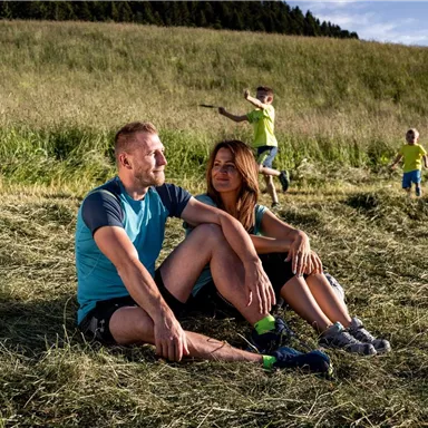 A couple sits in the tall grass and enjoys their time together. In the background, children play joyfully.