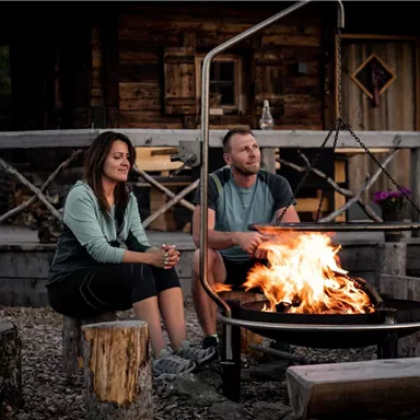 A couple sits relaxed by the campfire. In the background, a wooden barn is visible.