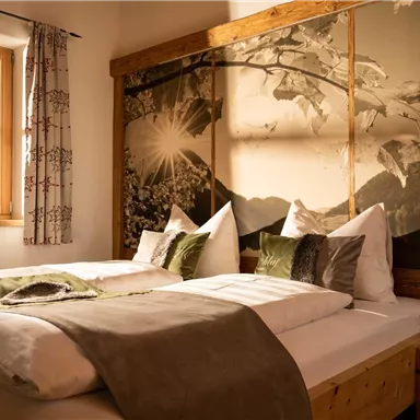 A cozy bedroom with two beds, decorated with cushions and a warm color palette. The window offers a view of nature and the wall is adorned with a beautiful wall mural.