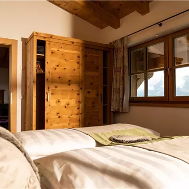 A cozy bedroom with two beds and wooden elements. Large windows offer a view of the surroundings and let in plenty of daylight.