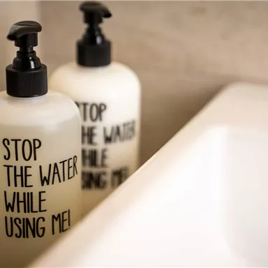 Two soap dispensers with the inscription "Stop the water while using me" are placed next to a sink. The dispensers have a simple design and convey a sustainable message.