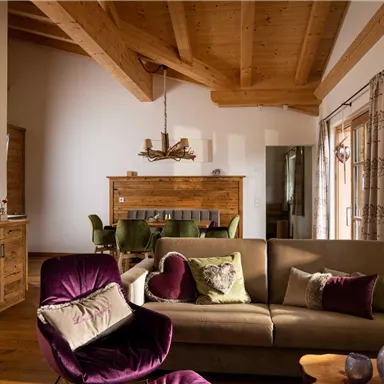 A cozy living room with a wooden ceiling and a large sofa. The decor is modern and inviting, with a dining area in the background.