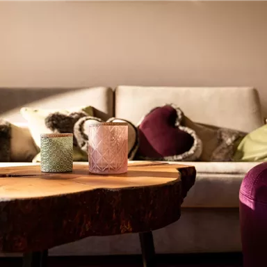 A cozy living room with a beige couch and colorful cushions. A wooden coffee table with two decorative glasses is in the foreground.