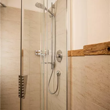 A modern shower enclosure with glass doors and an elegant showerhead. The walls are in neutral beige tones and feature a rustic wood accent.