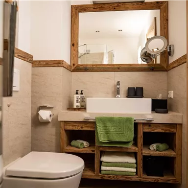 A modern bathroom with wood details and a large mirror. Care products and green towels are visible on the sink.