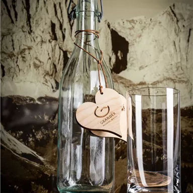 A stylish glass bottle with a wooden tag stands in front of a mountain landscape. To its right, a clear glass is placed on a wooden board.