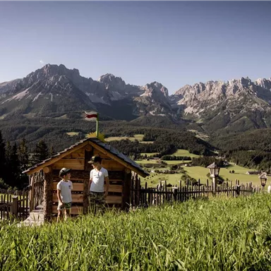 A picturesque landscape with tall mountains in the background and a small cabin in the foreground. Two people are standing on a green meadow.