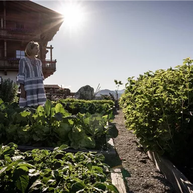 A woman is standing in a vegetable garden with lush plants. In the background, there is a wooden house and the sun is shining brightly.
