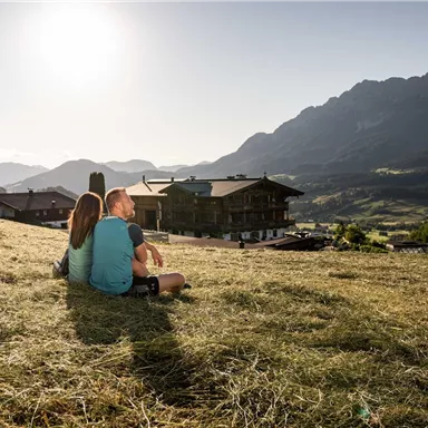 A couple sits on a meadow and enjoys the view of the mountains. The sun is shining, and the surroundings are peaceful and idyllic.