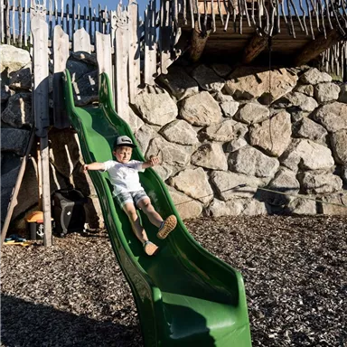A child is happily sliding down a green slide. In the background, you can see a stone wall and a wooden playhouse.