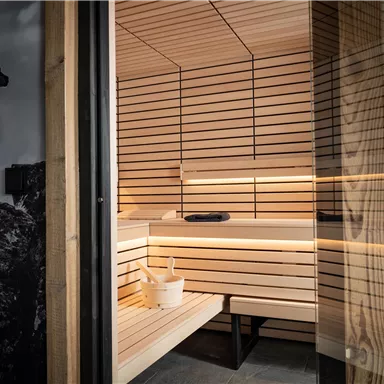 Eine moderne Sauna aus hellem Holz mit sanfter Beleuchtung. Im Vordergrund steht ein Eimer für Wasser und ein Becken, das Gemütlichkeit ausstrahlt.