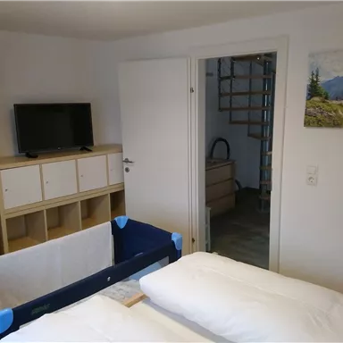 A simple bedroom with a TV, a wardrobe, and a children's bed. A picture of a mountain landscape hangs on the wall.