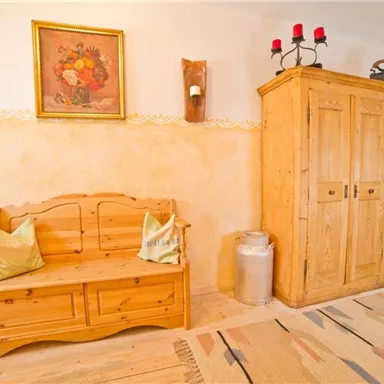 Ein gemütlicher Raum mit einer Holzbank und einem Holzschrank. An der Wand hängt ein Blumenbild und ein Tisch mit Blumen steht in der Ecke.