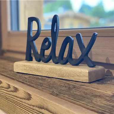 Ein dekoratives Schild mit dem Wort "Relax" aus schwarzem Material steht auf einem Holztisch. Im Hintergrund ist ein Fenster zu sehen, das einen natürlichen Lichtschein ermöglicht.