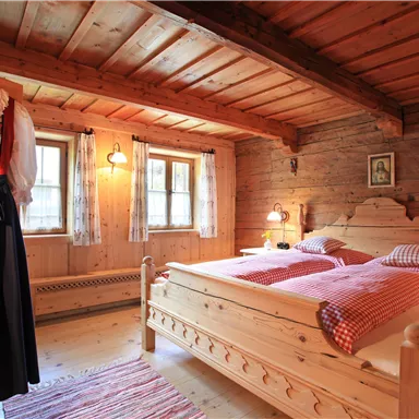 A cozy bedroom with wooden walls and a double bed. A traditional dress hangs on the wall and there are large windows with curtains.