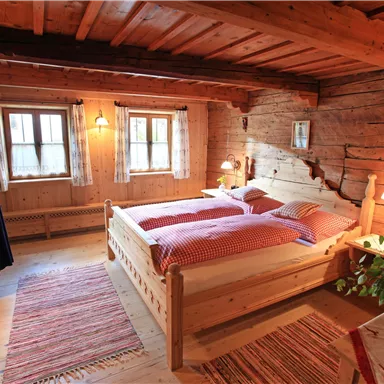 A cozy bedroom with a wooden bed and red bedding. The walls are made of wood, and there are windows that let in plenty of light.