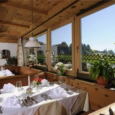 A cozy dining room with wooden furniture and beautifully set tables. Through the windows, one looks out onto a green landscape and blue sky.