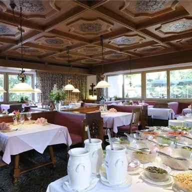 A cozy restaurant with wooden elements and warm lighting. Several tables are set for guests, and a buffet with a variety of dishes is visible.