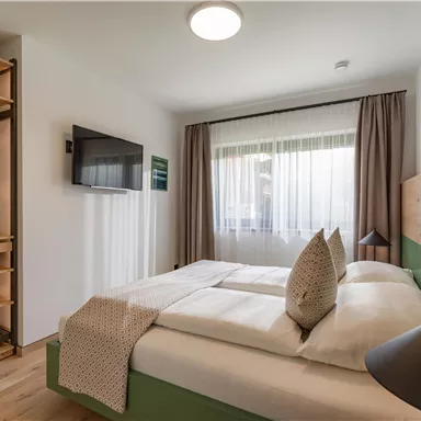 A modern bedroom with a double bed, two pillows, and a television on the wall. Large windows provide plenty of daylight and a bright atmosphere.