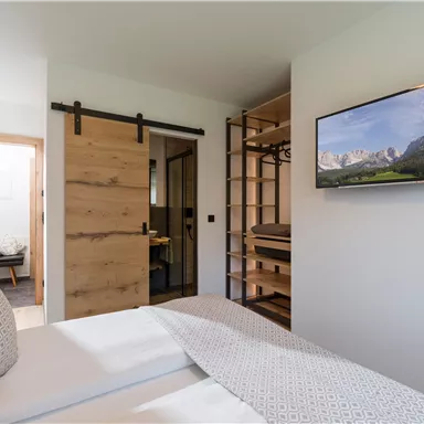 A modern bedroom with wooden details and a large window.  
On the wall, there is a television and a cozy bed.