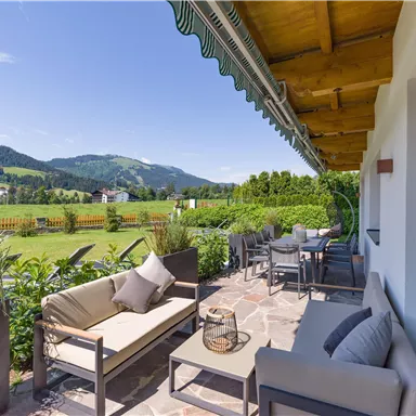 A cozy terrace with modern furniture and a view of the green landscape. In the background, there are gentle hills and a blue sky.