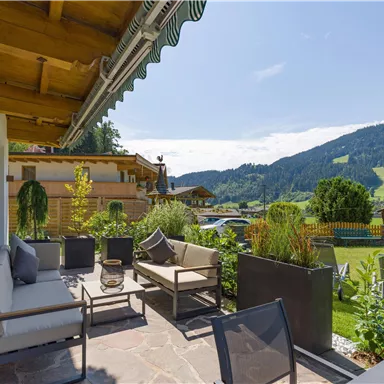 A cozy terrace with modern furniture, surrounded by green plants. In the background, majestic mountains and a blue sky are visible.