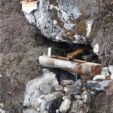 Eine kleine Nische in den Felsen mit einem Holzkreuz und einem Wasserlauf. Umgeben von Steinen und Holzstämmen.
