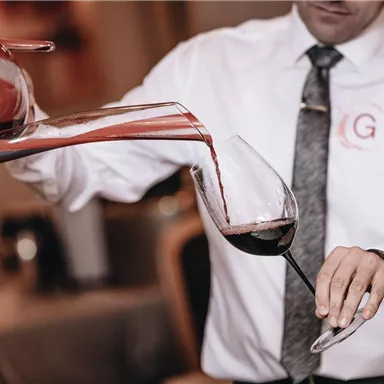 A waiter carefully pours red wine from a decanter into a glass. The background features an elegant restaurant atmosphere.