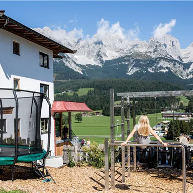 Ein idyllisches Haus mit einem Garten und Spielplatz, umgeben von majestätischen Bergen. Kinder spielen fröhlich auf der Schaukel und dem Trampolin.