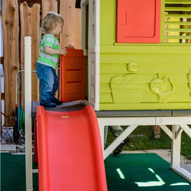 Ein kleines Kind spielt auf einer Rutsche vor einem bunten Spielhaus. Im Hintergrund sind ein Holzzaun und grünes Gras zu sehen.