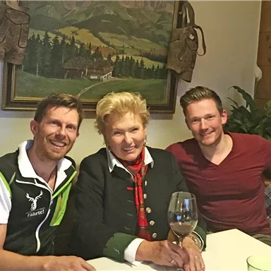 Three people are sitting at the table in a cozy environment. One person is holding a glass of wine and in the background, there is a picture of a landscape visible.