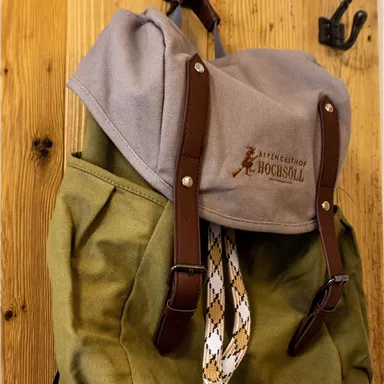 A green backpack with a gray lid and brown straps hangs on a hook. The backpack has a simple, rustic appearance.