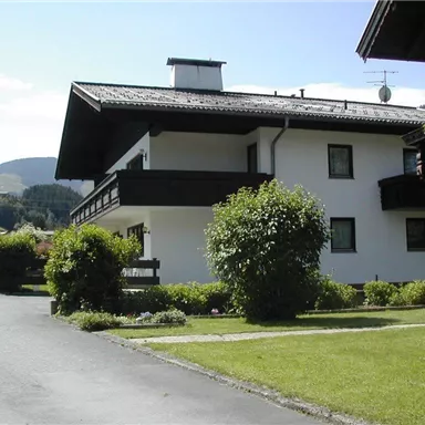 A quiet building in a rural setting with a well-maintained garden. In the background, gentle hills and a blue sky can be seen.
