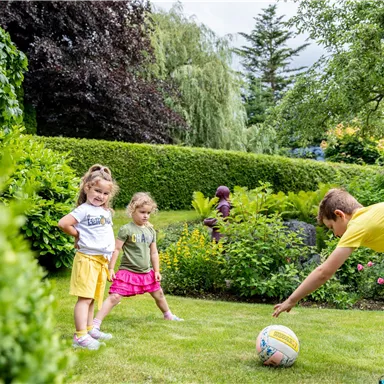 Two little girls and a boy are playing with a ball in a beautiful garden. Surrounded by green plants and trees, they are enjoying the sunny day.