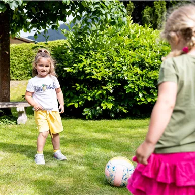 Two children are playing in the garden with a soccer ball. The girl in yellow is smiling, while the other girl in pink is ready to kick the ball.