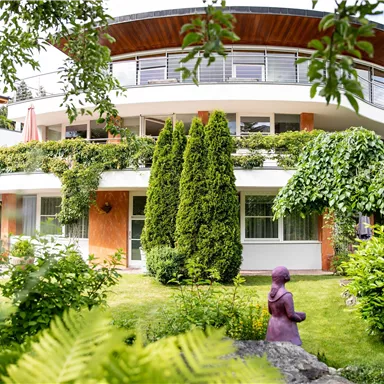 A modern building with a round balcony, surrounded by lush vegetation. In the foreground stands a purple statue in a well-kept garden.
