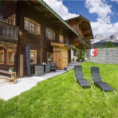Ein hübsches Chalet mit Holzfassade liegt in einer grünen Wiese. Zwei Liegen stehen zum Entspannen bereit, während die Berge im Hintergrund sichtbar sind.