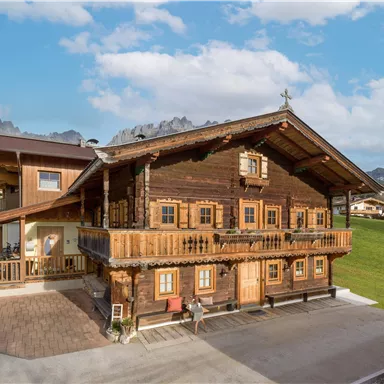Ein traditionelles Holzhaus mit einem Balkon und vielen Fenstern. Im Hintergrund sind grüne Wiesen und Berge sichtbar.