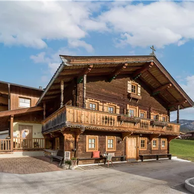 Ein traditionelles Holzhaus mit einem schönen Balkon und einer hellen Fassade. Im Hintergrund sind sanfte Hügel und ein blauer Himmel zu sehen.