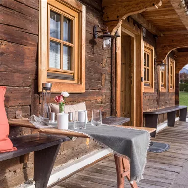 Ein gemütliches Holzhaus mit großen Fenstern und einer einladenden Veranda. Auf einem Tisch stehen Gläser und eine Vase mit Blumen, die eine freundliche Atmosphäre schaffen.
