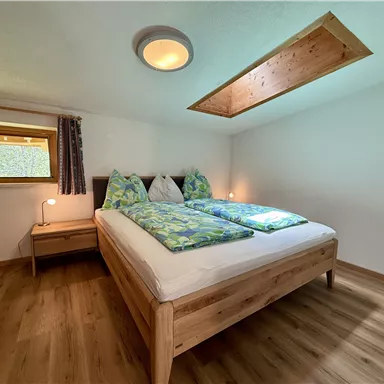 A cozy bedroom with a double bed and natural wooden furniture. There is a window that lets in daylight.