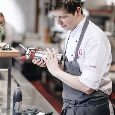 A chef in a modern kitchen is preparing a dish with a siphon. On the table, there is a dessert, and in the background, kitchen appliances are visible.