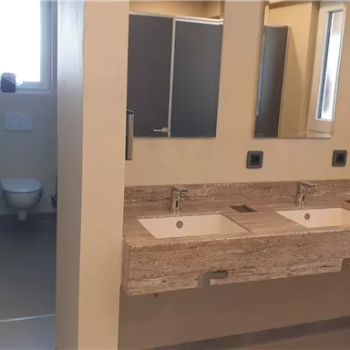 Ein modernes Badezimmer mit zwei Waschbecken und großen Spiegeln. Im Hintergrund sind getrennte Toilettenräume zu sehen.