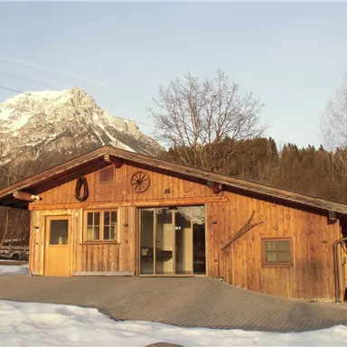 Ein Holzhaus mit großen Fenstern und einem schönen Berg im Hintergrund. Davor liegt Schnee und die Landschaft ist ruhig und idyllisch.
