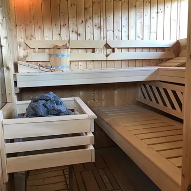Eine traditionelle Sauna aus Holz mit Sitzbänken und einem Aufgussbehälter. Die Wände sind aus hellem, poliertem Holz und es herrscht eine entspannende Atmosphäre.
