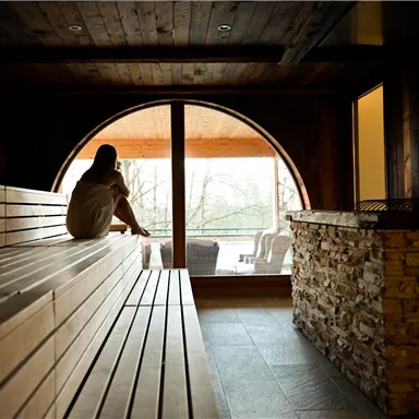 Eine ruhige Sauna mit Holzbank und sanfter Beleuchtung. Durch ein großes Fenster ist der Blick ins Freie zu sehen.